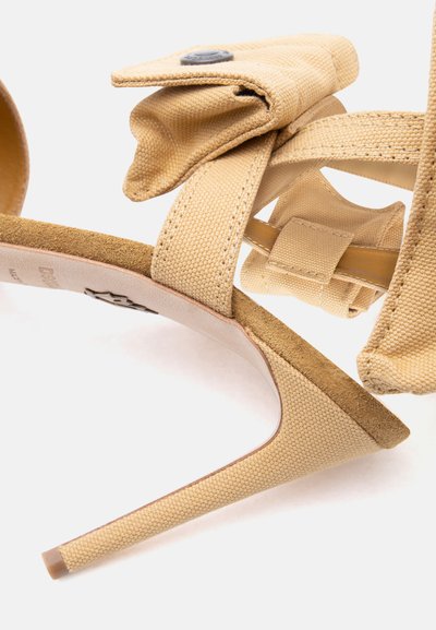 Μπεζ σανδάλι με ψηλό τακούνι, υφασμάτινα λουριά και μικρή προσαρτημένη θήκη που ασφαλίζει με κουμπί στον ιμάντα του αστραγάλου.