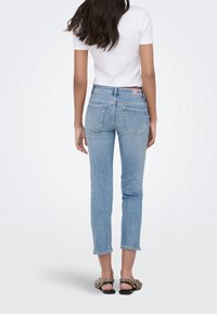 Femme aux longs cheveux foncés portant un jean court bleu clair, un haut blanc à manches courtes et des sandales noires et blanches à motifs, debout de dos.