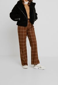 Musta tekoturkismantteli vetoketjullisilla taskuilla, ruskeat ruutukuvioiset korkeavyötäröiset housut ja valkoiset tennarit. Teksturoitu neulepaita alla.