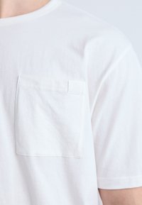 White cotton t-shirt with a round neckline, short sleeves, and a chest pocket featuring a small logo tag. Smooth texture and classic fit.