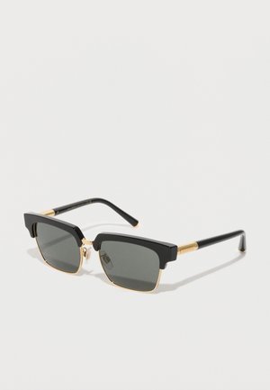 Lunettes de soleil rectangulaires noires avec des verres gris foncé, des accents en métal doré et des détails uniques en forme de sourcil sur la monture.