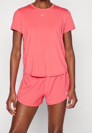 Osoba o ciemnej karnacji stojąca, ubrana w różowy sportowy t-shirt z krótkim rękawem i pasujące szorty z białymi logo Nike, ręce rozluźnione wzdłuż ciała.