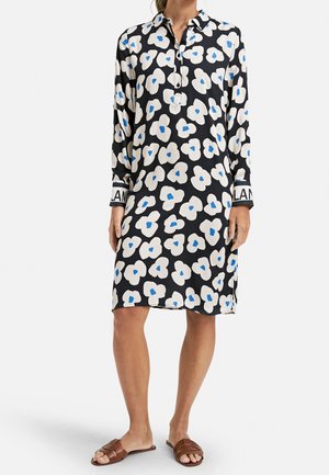 Frau trägt ein schwarzes Hemdkleid mit großen weißen Blumen mit blauen Zentren, langen Ärmeln und braunen flachen Sandalen.