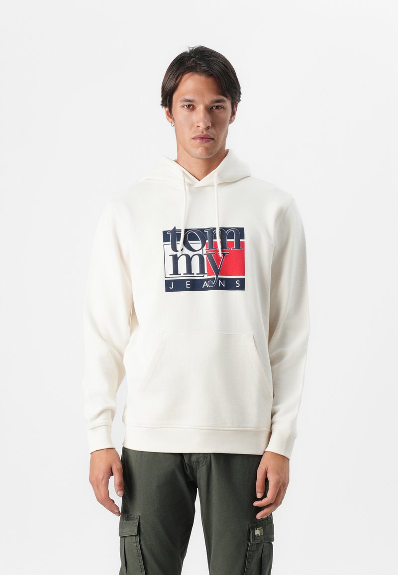 Krémová mikina s klokaní kapsou, s grafickým logem "tommy JEANS" v námořnické modré, červené a bílé barvě na přední straně. Měkká tkanina, nastavitelná šňůrka.