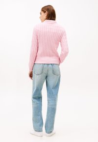 Rosa gerippter Strickpullover mit hohem Kragen, kombiniert mit hellblauen Jeans mit geradem Bein und weißen Turnschuhen. Minimalistischer Stil mit klaren Linien.