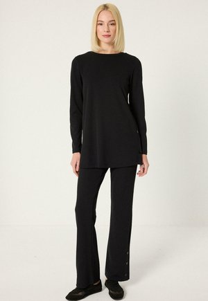 Szőke nő áll, fekete hosszú ujjú tunika felsőt és hozzáillő fekete bő szárú nadrágot, valamint fekete bebújós cipőt visel.