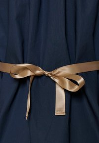 Sima textúrájú tengerészkék anyag, amely csomózott aranyszatén övvel díszített derékrészén, kontrasztot és eleganciát kölcsönözve.