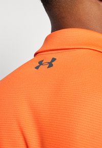 Polo naranja con textura acanalada, que presenta un logo negro de Under Armour en el cuello trasero. Diseño limpio, de manga corta.