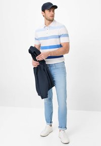Polo shirt à rayures blanches et bleues, jeans bleu clair et baskets blanches. Tient une veste bleue marine, porte une casquette bleue marine. Tenue décontractée.