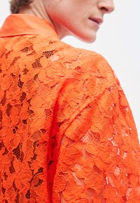 Oranje kanten blouse met een kraag, met bloemmotieven en een doorschijnende textuur. De nadruk ligt op fijne details en levendige kleur.