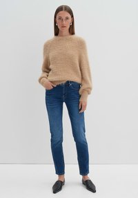 Beige fluffy gebreide trui met lange mouwen, gecombineerd met strakke donkerblauwe jeans. Zwarte platte instappers maken de outfit af. Eenvoudige achtergrond.