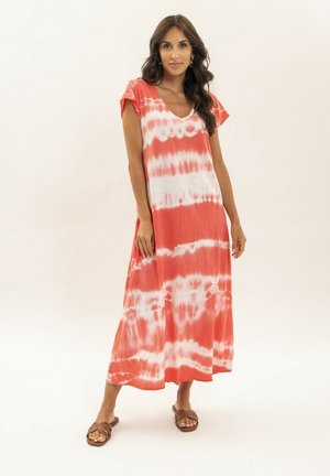 Femme debout, portant une robe maxi tie-dye corail et blanche à manches courtes et des sandales marron à enfiler, sur un fond uni.