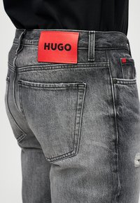 Sive traperice s stražnjim džepom i crvenom oznakom branda "HUGO" na pojasu, nošene s crnim topom.