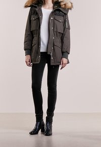 Olivově zelená parka s kapucí z umělého kožešiny, několika kapsami, stahovacími pasem a zipovým zapínáním. V kombinaci s černými skinny džíny a botami.