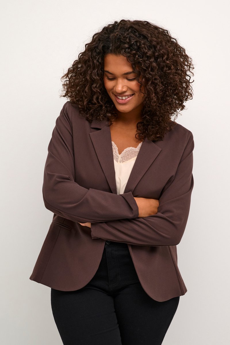 Blazer marron ajusté avec une texture lisse, fermeture à un seul bouton, col cranté et manches longues, porté sur un haut en dentelle léger.
