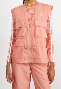 Kvinna i en korallrosa Adidas-väst över en mönstrad långärmad tröja med vita ränder och matchande byxor, framifrån, med avslappnade händer.