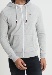 Světlounce šedá mikina na zip s kapucí se stahovací šňůrkou, předními kapsami a žebrovanými manžetami. Má kontrastní vnitřek kapuce a drobný detail s logem.