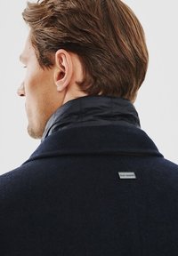 Granatowy płaszcz z wysokim kołnierzem, o miękkiej wełnianej teksturze i czarnym pikowanym wewnętrznym kołnierzu. Zawiera metalowy znacznik z tyłu.