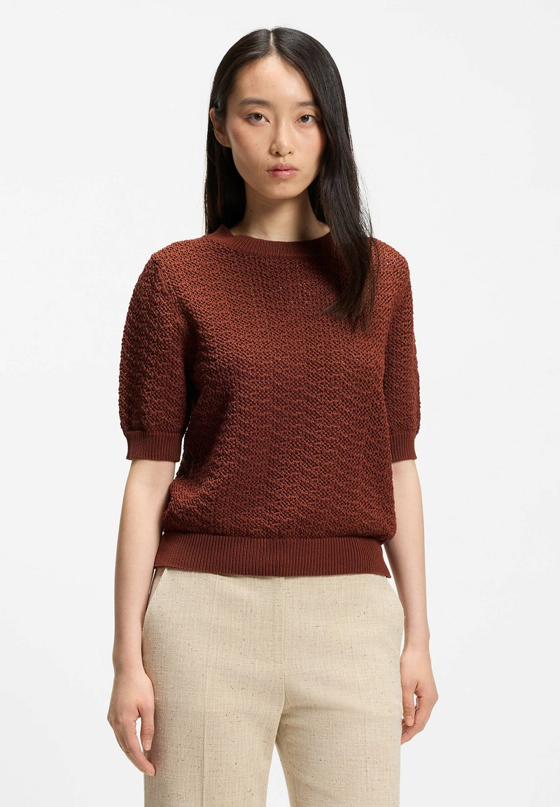 Femme aux longs cheveux foncés portant un pull texturé à manches courtes couleur rouille et un pantalon beige clair, debout devant un fond blanc uni.