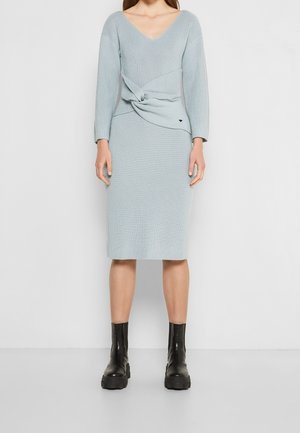 Donna che indossa un vestito lavorato a maglia azzurro chiaro con dettaglio intrecciato in vita e stivaletti neri con suola spessa, in piedi davanti a uno sfondo semplice.