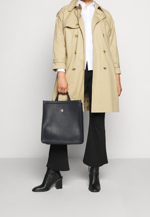Personne portant un trench-coat beige, une chemise blanche, un pantalon noir et des bottines à talons noires, tenant un grand sac fourre-tout noir par ses poignées.