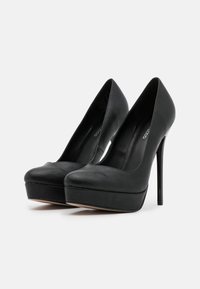 Svart høyhælte sko laget av glatt lær med rund tå og plattform, med en blank stiletthæl.