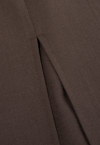 Bruine stof met een gestructureerd oppervlak, voorzien van een verticale naad en splesse. Het materiaal lijkt stevig en is geweven.