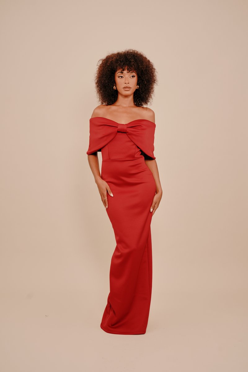 Žena s kučeravými vlasmi, oblečená v červenej off-shoulder róbe s veľkým mašľovým detailom, stojí na pozadí v jemnej béžovej farbe.