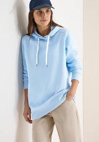 Jeune femme portant un sweat à capuche bleu clair et un pantalon beige, debout contre un mur blanc avec une main dans la poche et une casquette bleue sur la tête.