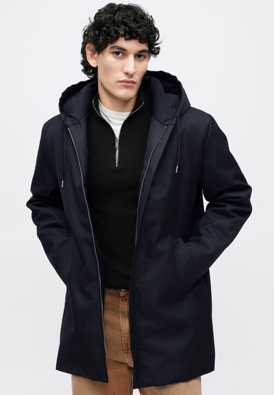 Tumesinine kapuutsiga mantel, millel on esikinnis, kaks küljetaskut ja lõdva lõikega istuvus. Kantud musta kampsuni ja pruunide pükstega. Sile kangapind.