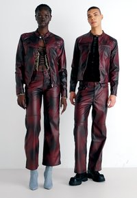 Tamno burgundska kožna jakna i hlače, s glatkom teksturom i prilagođenim dizajnom. Model nosi plave čizmice do gležnjeva, s vidljivim šavnim detaljima.