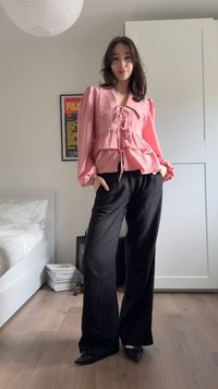 Nő áll egy szobában, rózsaszín hosszú ujjú fodros blúzt és fekete, széles szárú nadrágot visel, kezei a zsebében, a kamerába mosolyog.