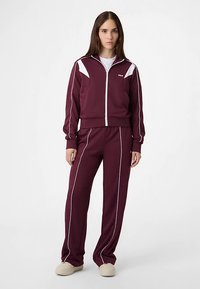 Punaruskea verkkopuku, jossa valkoiset korostusviivat, vetoketjullinen takki ja kapealahkeiset housut. Pehmeästä materiaalista valmistettu, suunniteltu mukavuutta varten.