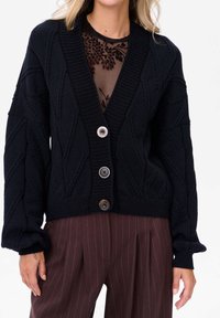 Czarny dziergany kardigan z głębokim dekoltem w serek, z teksturowanymi wzorami w diamenty i zapięciem na guziki. Długie, bufiaste rękawy i ściągacz przy dole.