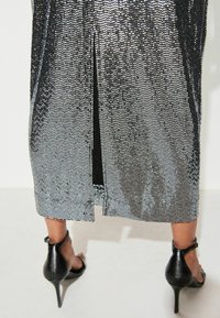 Onderbenen in zwarte hoge hakken met enkelbandjes onder een midi-jurk met zilveren sequins en een split aan de achterkant.
