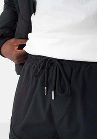 Zwarte drawstring shorts met een elastische tailleband, voorzien van zilveren aglets, gecombineerd met een wit shirt en een zwarte jas. Soepele textuur.