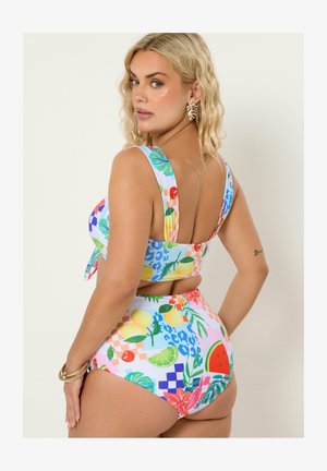 Kvinde med blondt hår iført en farverig højttilpasset bikini med frugt- og blomsterprint, guldfarvede øreringe formet som blade samt guldbøjler, der kigger over skulderen.