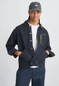 Νεαρός με πλεξούδες που φοράει ένα σκούρο καπέλο, ναυτικό σακάκι με μπροστινές τσέπες, λευκό πουκάμισο και ναυτικά παντελόνια, με τα χέρια στις τσέπες, χαμογελάει.