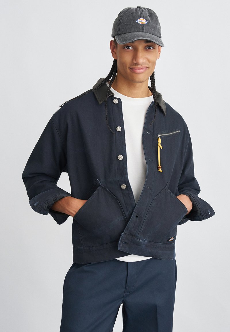 Νεαρός με πλεξούδες που φοράει ένα σκούρο καπέλο, ναυτικό σακάκι με μπροστινές τσέπες, λευκό πουκάμισο και ναυτικά παντελόνια, με τα χέρια στις τσέπες, χαμογελάει.