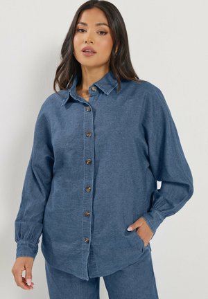 Mujer con una camisa de pana azul abotonada y pantalones a juego, posando con una mano en el bolsillo contra un fondo liso.