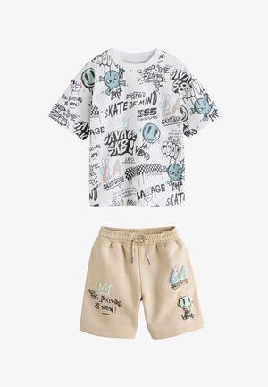 Dreng står med hænderne i lommerne på sine korte bukser og har en hvid t-shirt med mønster på, beige korte bukser med grafik, hvide sokker og hvide sneakers med sorte striber.