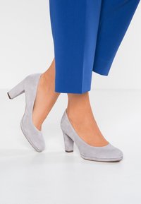 Chaussures à talons hauts en suede gris avec une pointe arrondie et un talon épais. Portées avec un pantalon cropped bleu, mettant en valeur une texture lisse et une silhouette épurée.