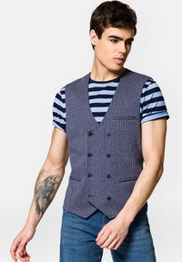 Niebiesko-biała kamizelka w wzór houndstooth z dekoltem w serek, podwójnymi guzikami i jedną kieszenią na piersi, noszona na paskowaną koszulkę.