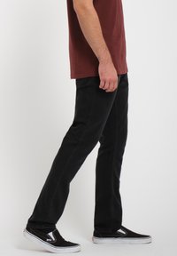 Black denim pants with straight cut, visible stitching details, worn with a maroon t-shirt and black slip-on sneakers.