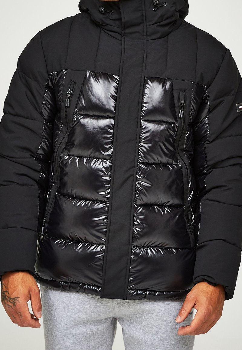 Svart puffjacka med en kombination av matta och glansiga material, vertikal sömnad, två zip-fickor på bröstet och en justerbar huva.