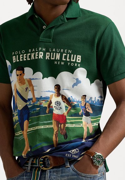 Camisa polo verde con un gráfico vibrante de tres corredores contra un paisaje. El diseño incluye texto del logotipo y un cuello clásico.