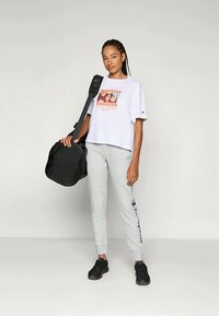 Kvinde med fletninger står og holder en sort træningspose, iført en hvid t-shirt med grafik, grå sweatpants og sorte sneakers mod en ensfarvet baggrund.