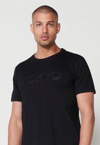 Melna īsām piedurknēm T-krekls ar "TOKYO" tekstu diskrētā printā. Mīksts audums, klasiskā apaļā kakla dizains. Vienkāršs un daudzpusīgs stils.