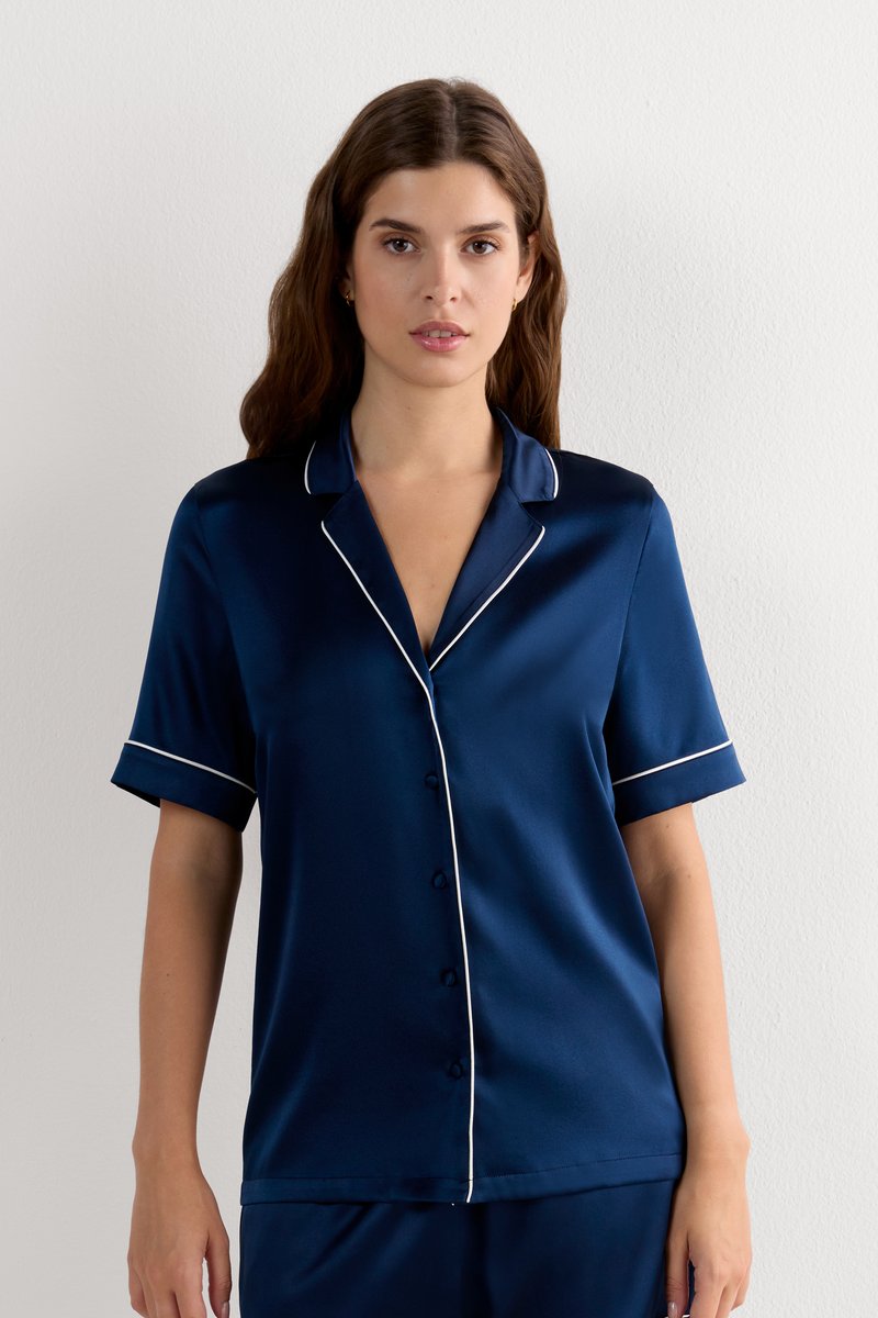 Vrouw met lang bruin haar die een marineblauw satijnen kortärmelig overhemd met witte bies draagt tegen een effen muur.