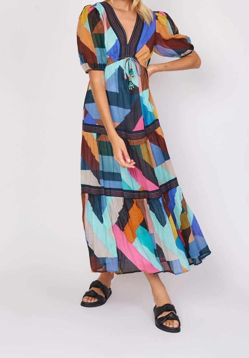 Robe longue multicolore avec un décolleté en V et des manches bouffantes, présentant des motifs géométriques et un lien orné à la taille. Portée avec des sandales noires.